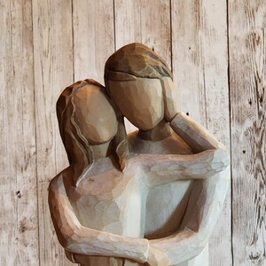 May include: A wooden sculpture of a couple embracing. The man is on the right and the woman is on the left. They are both carved in a simple, stylized way. The sculpture is painted in shades of brown and white.