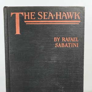 May include: A black book with the title "THE SEA-HAWK" in red lettering. The author, "BY RAFAEL SABATINI", is printed in red below the title.