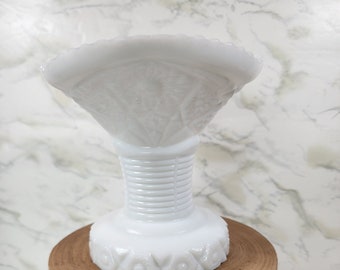 May include: White milk glass vase with a scalloped top and a ribbed stem. The vase is sitting on a wooden stand with black metal legs.