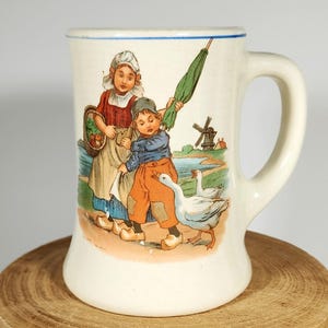 May include: A white ceramic mug with a blue rim and handle. The mug features a colorful illustration of two children, a girl with a basket of vegetables and a boy holding an umbrella, walking with two white geese. The scene is set in a rural landscape with a windmill in the background.