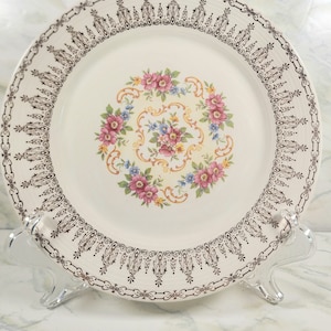 May include: A white ceramic plate with a gold rim and a floral design in the center. The flowers are pink, blue, and yellow. The plate is on a white marble surface.