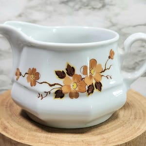 May include: White ceramic creamer with a floral design featuring brown leaves and orange flowers. The creamer has a rounded body and a handle.