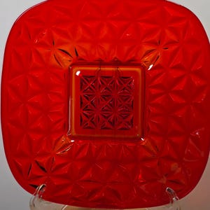 May include: A square, red glass plate with a textured, geometric pattern. The center of the plate features a smaller square with a crosshatch design. The plate is displayed on a clear stand.