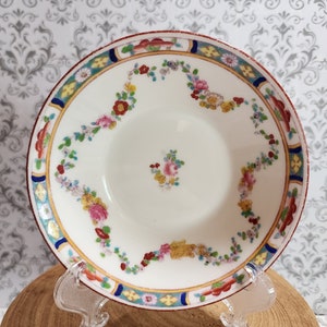 May include: A white porcelain saucer with a colorful floral pattern. The saucer has a red, blue, and gold border with a delicate floral design in the center.