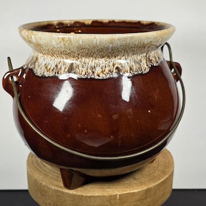 May include: A brown ceramic pot with a cream-colored rim and a metal handle. The pot has a glossy finish and sits on a wooden base. The pot is likely used for cooking or decoration.