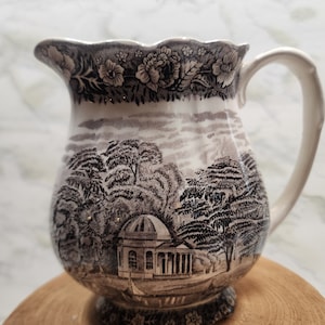 May include: A vintage ceramic pitcher with a black and white landscape design. The pitcher features a scalloped rim with floral detailing and a curved handle. The landscape depicts trees and a domed building.