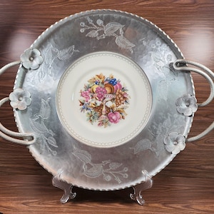 May include: A silver serving tray with two handles and a raised rim. The tray has a floral design and a white porcelain plate in the center with a floral design and fruit.