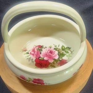 May include: A white ceramic bowl with a handle, featuring a floral design of pink and red roses. The bowl has a glossy finish and is decorated with a delicate gold trim.