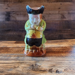 May include: A ceramic pitcher shaped like a man wearing a green jacket, black pants, and a black hat. The man has a white face with painted details and a brown beard.