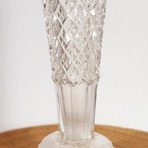May include: A clear glass vase with a diamond pattern. The vase has a round base and a tall, narrow neck.