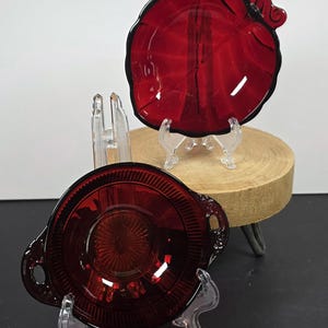 May include: Two vintage ruby red glass serving dishes. One is a round bowl with handles and a textured pattern. The other is a leaf-shaped dish with a black trim. Both are displayed on clear stands.