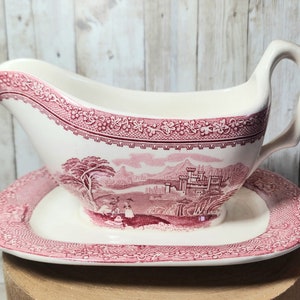 May include: A white ceramic gravy boat with a pink floral design and a matching saucer. The gravy boat has a handle and a spout. The design features a landscape with a castle and trees.