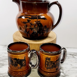 May include: A set of brown ceramic mugs and a pitcher with a black and white illustration of a historical scene. The mugs have a silver rim and handle.