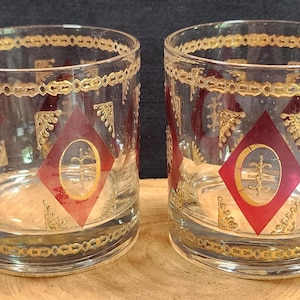 May include: Two clear glass tumblers with gold and red geometric designs. The glasses have a gold rim and a gold band around the middle.