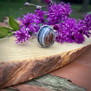 May include: A silver ring with a large, oval, striped gemstone in shades of brown, white, and blue. The gemstone is set in a twisted rope-like bezel. The ring is displayed on a wooden surface with purple flowers.