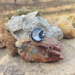 May include: A silver ring with a crescent moon design featuring a white gemstone and a small star. The ring is set on a rock.