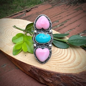 May include: A silver ring featuring a central oval turquoise cabochon flanked by two pink heart-shaped stones. The setting is detailed with twisted wire borders and small silver spheres, creating a vertical, symmetrical design on the band.