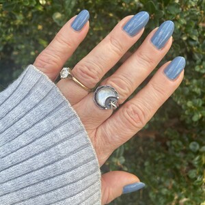 May include: A hand with light blue nail polish is wearing a silver ring with a white moon and a star design. The ring is on the middle finger.