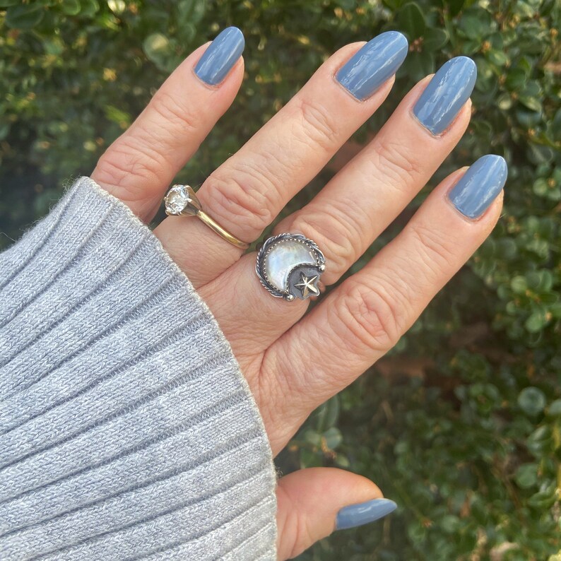 May include: A hand with light blue nail polish is wearing a silver ring with a white moon and a star design. The ring is on the middle finger.