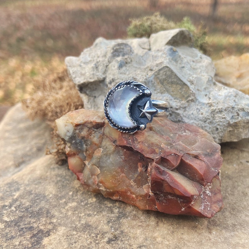 May include: A silver ring with a crescent moon design featuring a white iridescent stone and a small star. The ring is set on a rock.