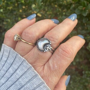 May include: A silver ring with a crescent moon and star design. The moon is made of white mother of pearl. The ring is worn on a finger with blue nail polish.