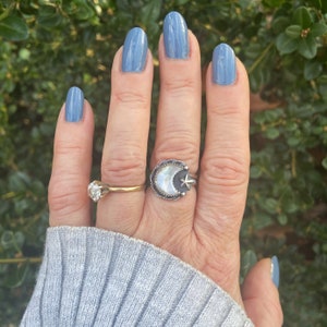 May include: A hand with light blue nail polish is wearing a silver ring with a white crescent moon and a star design. The ring is on the middle finger of the hand. The hand is also wearing a gold band on the ring finger.