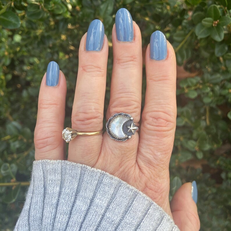 May include: A hand with light blue nail polish is wearing a silver ring with a white crescent moon and a star design. The ring is on the middle finger of the hand. The hand is also wearing a gold band on the ring finger.