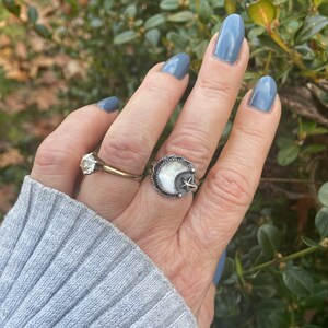 May include: A silver ring with a white mother-of-pearl crescent moon and a star design. The ring is on a person's finger.