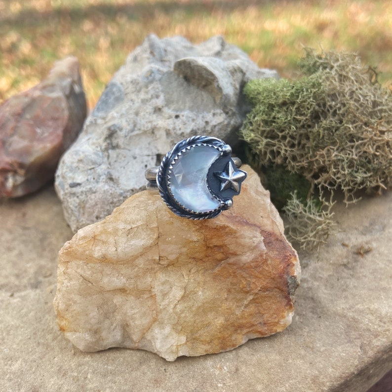 May include: A silver ring with a crescent moon design featuring a white iridescent stone and a small star accent.