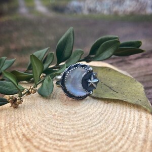 May include: A silver ring featuring a crescent moon with a white iridescent stone and a small star accent.