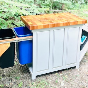 May include: A white kitchen island with a butcher block countertop and pull-out trash and recycling bins. The island has a gray painted finish and a wood top.