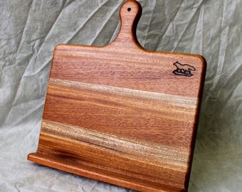May include: A wooden cutting board with a handle and a raised edge. The board has a natural wood grain and is stained a reddish brown color. The board has a small, black, stylized animal design on the top right corner.