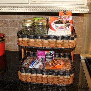 May include: Two-tiered wicker basket set with black accents. The top tier holds jars of tea, a bag of coffee, and a bag of marshmallows. The bottom tier holds a bag of bread and a box of granola bars. The text on the coffee bag reads "Dunkin' Donuts Original Blend".