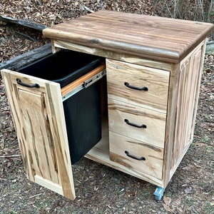 May include: A wooden kitchen island with a butcher block countertop, three drawers, and a pull-out trash bin. The island is on wheels and has a natural wood finish.