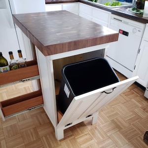 May include: A white kitchen island with a dark wood butcher block countertop. The island has a pull-out trash can and two pull-out shelves for storage. The island is on a hardwood floor.
