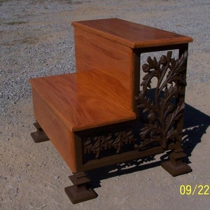 May include: Two-step wooden stool with a decorative iron scrollwork design featuring leaves and acorns. The stool has a dark brown finish and is supported by four decorative iron feet.