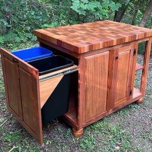 May include: A wooden kitchen island with a butcher block countertop and a pull-out trash bin system. The island has two cabinet doors and a shelf. The trash bins are black and blue.