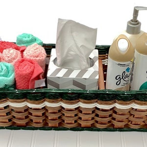 May include: A woven basket with a green and white trim contains rolled hand towels, a box of tissues, and two bottles of Glade air freshener.