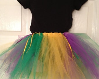 May include: A black short-sleeved bodysuit paired with a layered tutu. The tutu features green, yellow, and purple tulle, with yellow and purple ribbons. The outfit is displayed on a mannequin.
