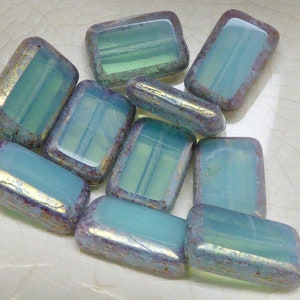 Peut inclure: Collection de perles rectangulaires bleu clair et opalescentes. Les perles ont une bordure métallique dorée et violette. Les perles sont de tailles et de formes variées, avec un bord légèrement arrondi.
