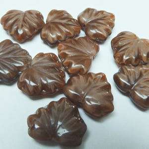May include: A selection of brown, leaf-shaped beads. Each bead features a detailed, veined design and a glossy finish. The beads are scattered on a white surface, highlighting their intricate details.
