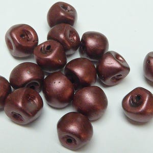 May include: A selection of maroon-coloured, irregularly shaped beads. Each bead has a hole through it, and they have a slight metallic sheen. The beads are scattered on a white surface, ideal for jewellery making or crafts.