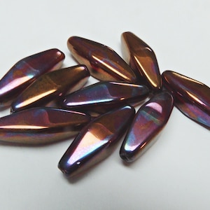May include: A collection of iridescent, oblong beads. The beads have a dark, reddish-brown base colour with shimmering highlights of purple, blue, and gold. The beads are scattered on a white surface, creating a visually appealing display.