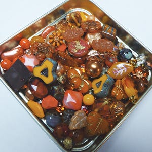 May include: A metal tray filled with a variety of orange, brown, and clear glass beads. The beads are in different shapes, including leaves, hearts, and squares.
