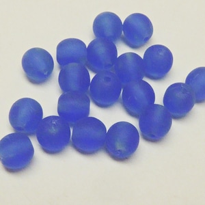 Peut inclure: Un ensemble de perles de verre bleu cobalt mat. Les perles sont rondes avec une forme légèrement aplatie et un aspect givré. Elles sont dispersées sur une surface blanche, adaptées à la fabrication de bijoux ou aux projets d'artisanat.