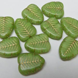 May include: A collection of green leaf-shaped beads with gold vein detailing. The beads are arranged on a white surface. These decorative beads are suitable for jewellery making and craft projects.