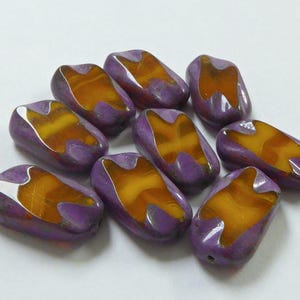 May include: A collection of nine rectangular glass beads featuring a smooth, opaque purple exterior with a central, translucent amber-coloured inlay. Each bead has a distinct, irregular cutout design on the top and bottom edges, revealing the amber glass.