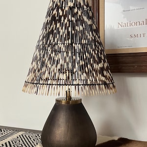 May include: A table lamp featuring a conical shade constructed from numerous black and white porcupine quills arranged in a vertical pattern. The lamp base is a smooth, dark brown, teardrop-shaped vessel with a metallic gold-toned neck fixture.