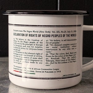 Peut inclure: Une tasse en &eacute;mail blanc avec un bord et une poign&eacute;e noirs. La tasse pr&eacute;sente une impression noir et blanc d'une d&eacute;claration des droits des peuples noirs du monde, publi&eacute;e en 1926 par le journal The Negro World. Le texte comprend la phrase "Ethiopie, Terre de nos P&egrave;res, etc., sera l'hymne de la race noire."