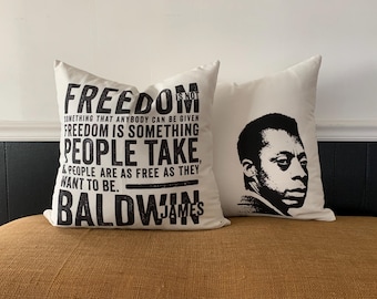 May include: Two white throw pillows with black text and a black and white portrait of James Baldwin. The text on one pillow reads "FREEDOM IS NOT SOMETHING THAT ANYBODY CAN BE GIVEN FREEDOM IS SOMETHING PEOPLE TAKE & PEOPLE ARE AS FREE AS THEY WANT TO BE. BALDWIN JAMES".